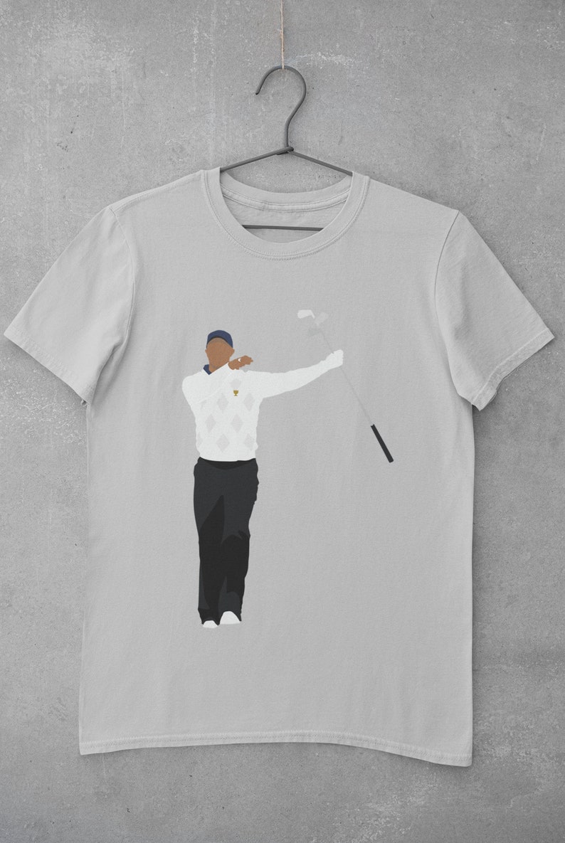May include: Light gray t-shirt featuring a minimalist illustration of a golfer in a white sweater with a diamond pattern, black pants, and a blue cap. The golfer is holding a golf club. The shirt is hanging on a metal hanger.