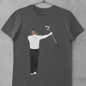 May include: Dark gray t-shirt featuring a stylized illustration of a golfer in a white sweater with a diamond pattern, black pants, and a blue cap. The golfer is holding golf clubs. The shirt is hanging on a metal hanger.