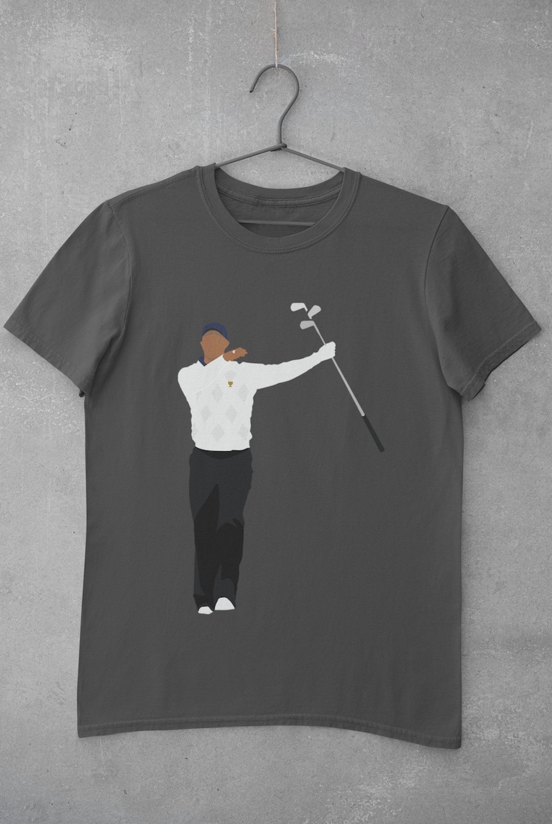May include: Dark gray t-shirt featuring a stylized illustration of a golfer in a white sweater with a diamond pattern, black pants, and a blue cap. The golfer is holding golf clubs. The shirt is hanging on a metal hanger.