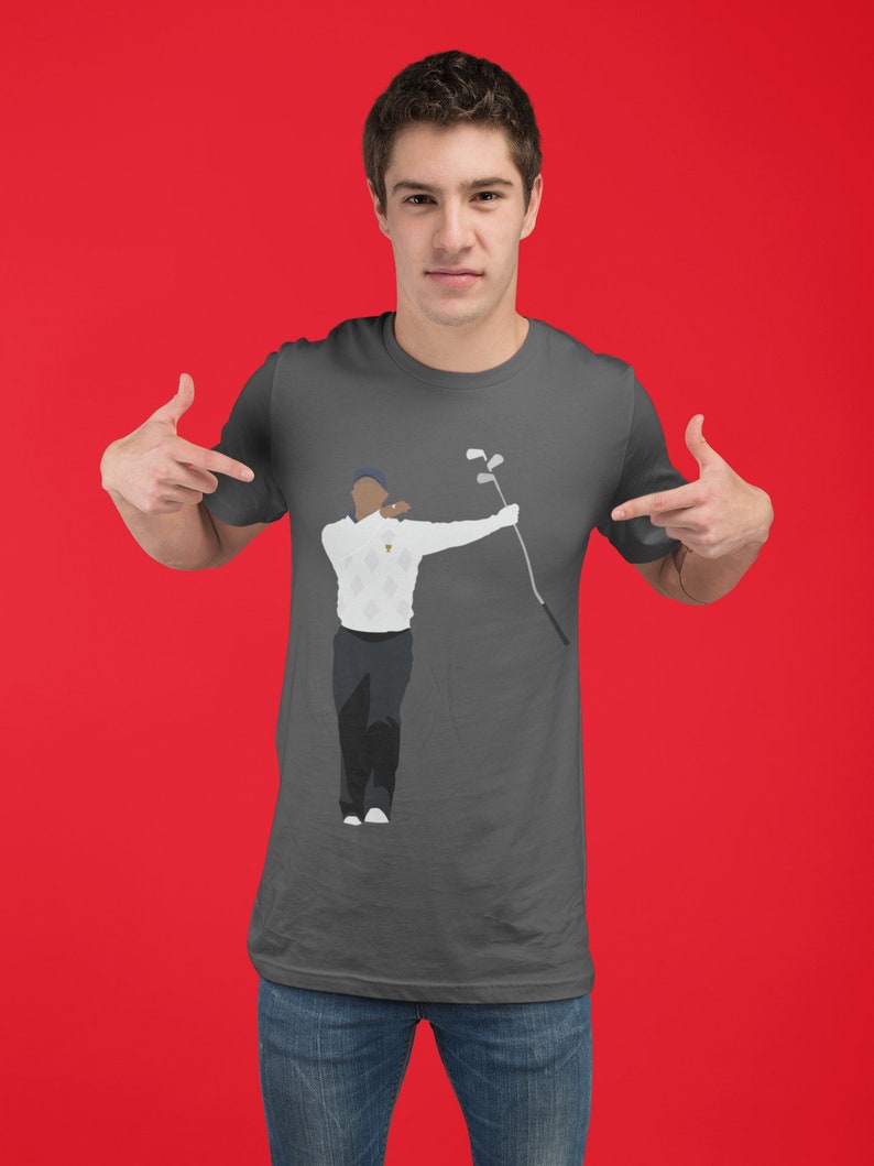 May include: A charcoal gray t-shirt featuring a graphic of a golfer in a white sweater and black pants, holding a golf club. The golfer is depicted in a mid-swing pose. The background is a solid red.