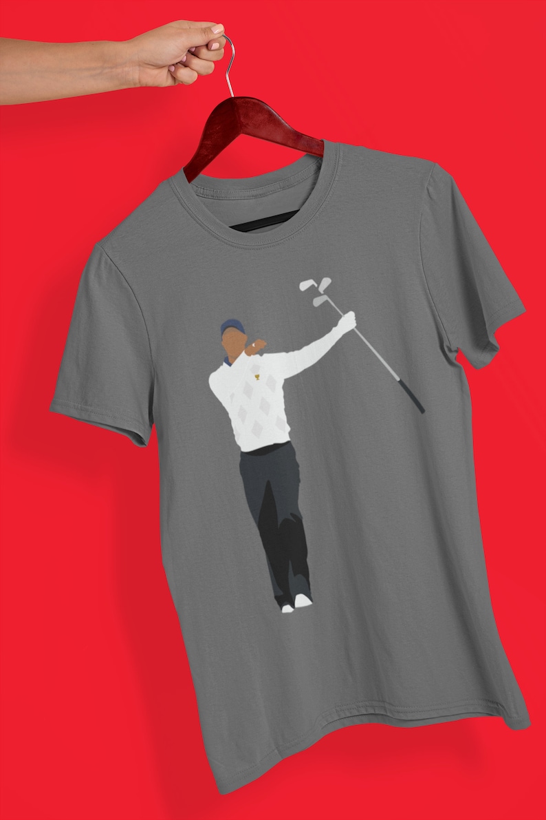 May include: Gray t-shirt featuring a graphic of a golfer in a white sweater with a diamond pattern, dark pants, and a blue cap. The golfer is mid-swing, holding a golf club. The shirt is hanging on a wooden hanger against a red background.