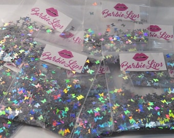 Könnte beinhalten: Mehrere kleine, transparente Plastiktüten gefüllt mit schimmerndem, glitzerndem Konfetti in Form von Schmetterlingen. Jede Tüte trägt ein Etikett mit der Aufschrift "Barbie Lips" und einem pinkfarbenen Lippenmotiv.