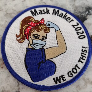 May include: A white embroidered patch with a blue border. The patch features a woman with brown hair wearing a blue shirt and a face mask. The text "Mask Maker 2020 We Got This!" is embroidered around the woman.