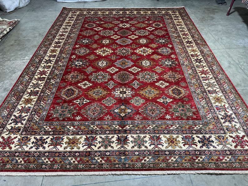 May include: A red, cream, and blue patterned rug with a geometric design. The rug has a fringe border.