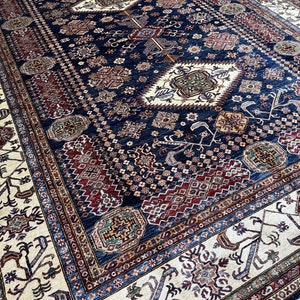 May include: A large, rectangular rug with an intricate, geometric pattern. The rug features a deep navy blue central field with cream, burgundy, and touches of green and gold. The border is cream with detailed designs. The rug is likely made of wool.