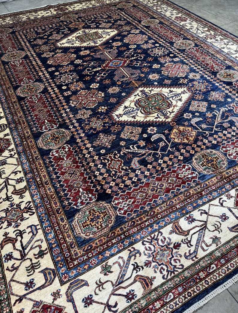 May include: A large, rectangular rug with an intricate, geometric pattern. The rug features a deep navy blue central field with cream, burgundy, and touches of green and gold. The border is cream with detailed designs. The rug is likely made of wool.