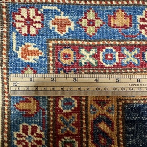 May include: Close-up of a section of a patterned rug with a ruler. The rug features a blue, red, and beige color scheme with floral and geometric designs. The ruler shows measurements in inches and centimeters.
