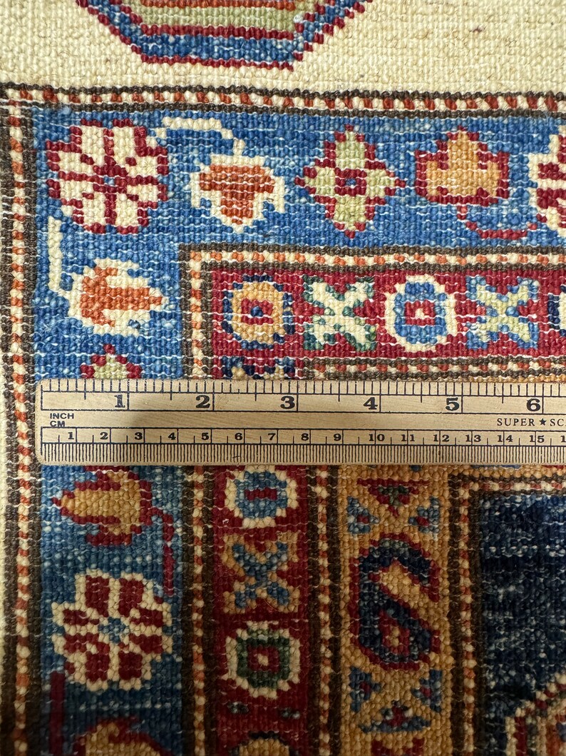 May include: Close-up of a section of a patterned rug with a ruler. The rug features a blue, red, and beige color scheme with floral and geometric designs. The ruler shows measurements in inches and centimeters.