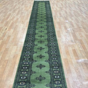 May include: A green runner rug featuring a central pattern of diamond shapes and floral designs. The rug has a dark border with a repeating pattern of white and black shapes. The rug is placed on a light-coloured wooden floor.