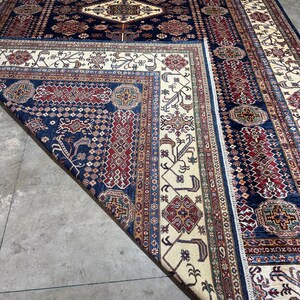 May include: A large, rectangular area rug with a traditional design. The rug features a navy blue field with a central diamond pattern and intricate borders in cream, red, and brown. The rug is partially folded, revealing the detailed pattern.
