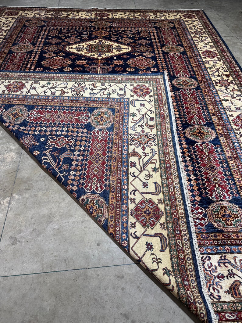 May include: A large, rectangular area rug with a traditional design. The rug features a navy blue field with a central diamond pattern and intricate borders in cream, red, and brown. The rug is partially folded, revealing the detailed pattern.