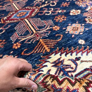May include: Close-up of a section of a blue and beige patterned rug. The rug features intricate geometric designs in shades of red, orange, and cream. The rug's texture is visible, with a hand holding up a portion of the rug.