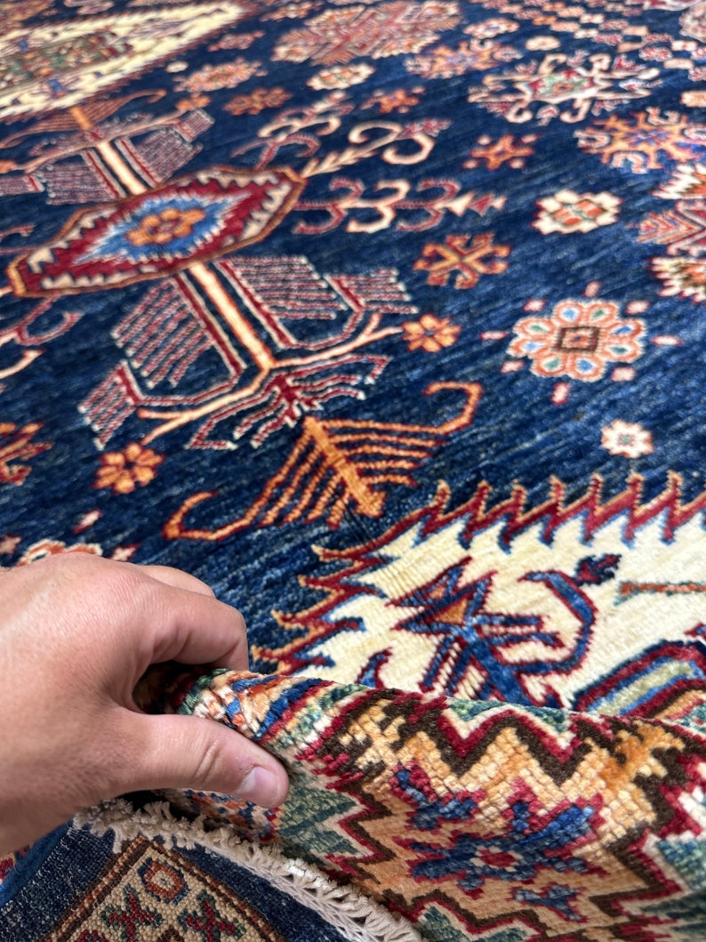 May include: Close-up of a section of a blue and beige patterned rug. The rug features intricate geometric designs in shades of red, orange, and cream. The rug's texture is visible, with a hand holding up a portion of the rug.