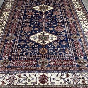 May include: A large, rectangular area rug with an intricate, geometric pattern. The rug features a deep navy blue central field with cream, red, and gold accents. The border is cream with repeating patterns. The rug is likely made of wool.