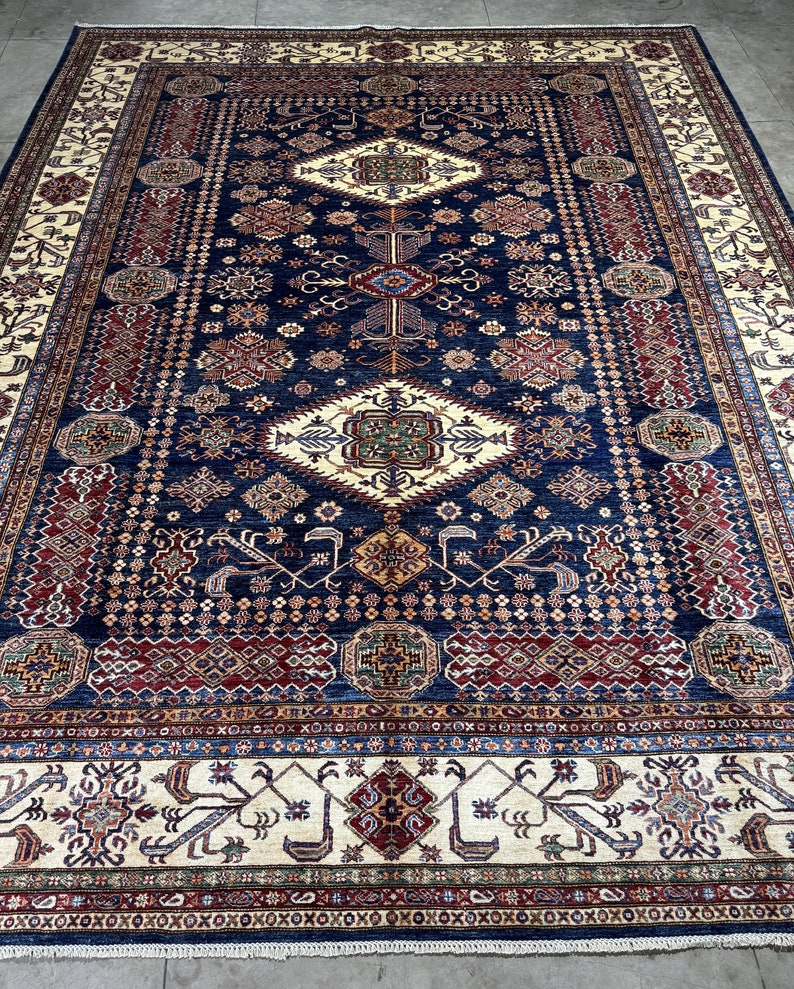 May include: A large, rectangular area rug with an intricate, geometric pattern. The rug features a deep navy blue central field with cream, red, and gold accents. The border is cream with repeating patterns. The rug is likely made of wool.
