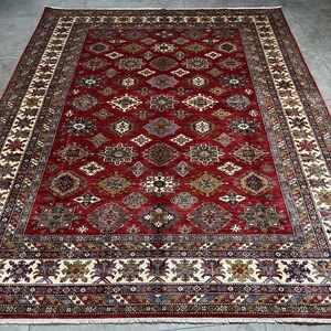 May include: A red, beige, and blue patterned rug with a geometric design. The rug has a detailed border with a repeating pattern.