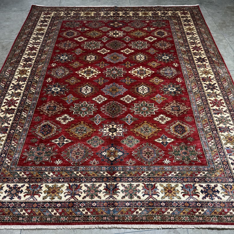 May include: A red, beige, and blue patterned rug with a geometric design. The rug has a detailed border with a repeating pattern.