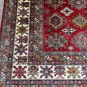 May include: A red, blue, and cream colored rug with intricate geometric patterns. The rug has a fringe border.