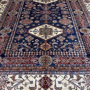 May include: A large, rectangular rug with an intricate, geometric pattern. The rug features a deep navy blue central field with a complex arrangement of red, beige, and gold motifs. The border is a lighter beige with repeating patterns.