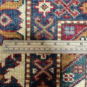 May include: Close-up of a beige rug with intricate patterns in shades of red, blue, and gold. A ruler is placed on the rug, showing the detail of the weave.