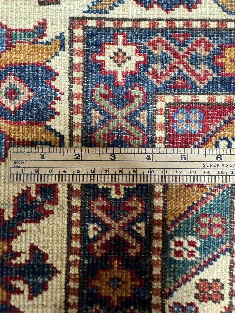 May include: Close-up of a beige rug with intricate patterns in shades of red, blue, and gold. A ruler is placed on the rug, showing the detail of the weave.