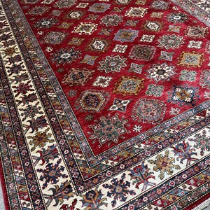 May include: A red, beige, and blue patterned rug with a geometric design. The rug has a fringe border.
