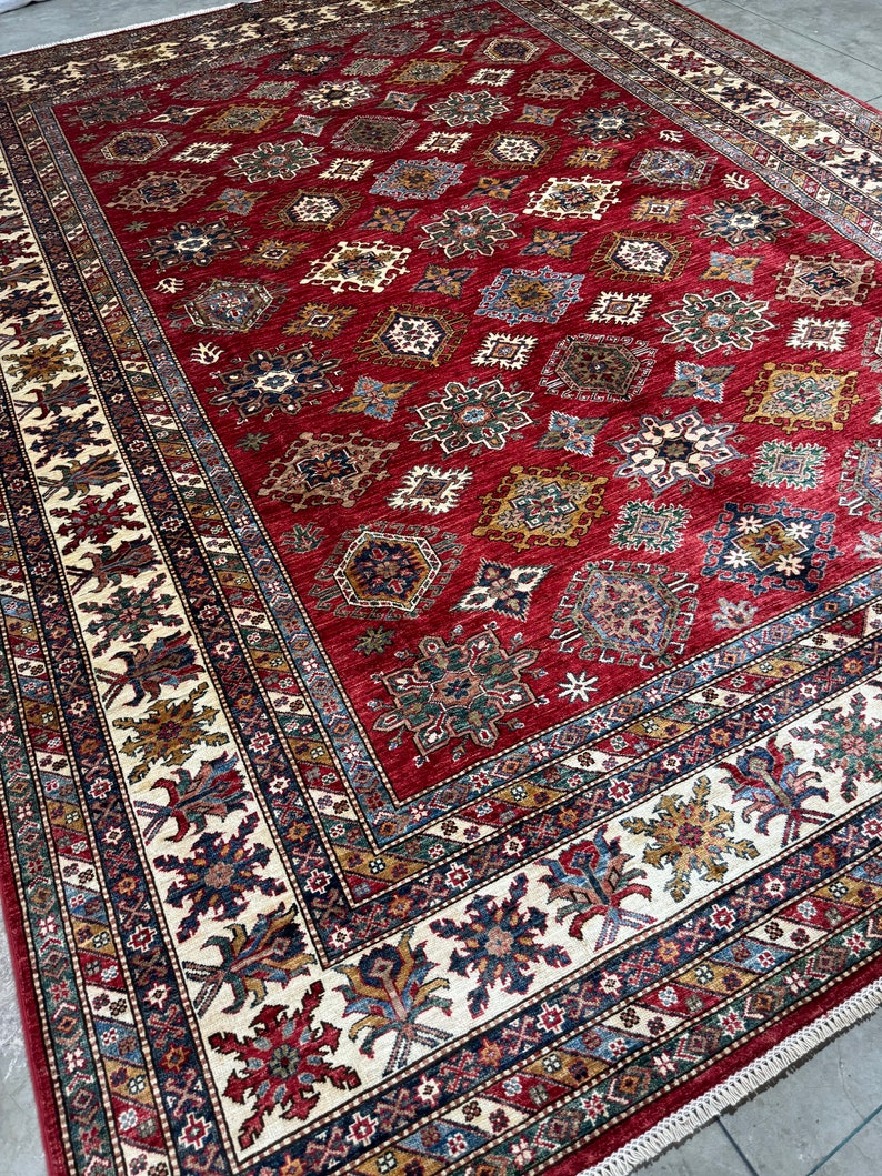May include: A red, beige, and blue patterned rug with a geometric design. The rug has a fringe border.