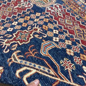 May include: Close-up of a section of a patterned rug. The rug features a deep blue field with intricate geometric and floral designs in red, beige, and gold. The detailed patterns create a rich, textured appearance, showcasing traditional craftsmanship.