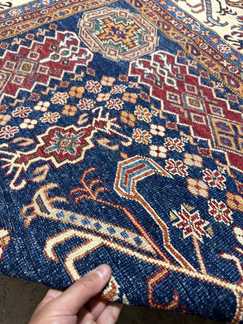 May include: Close-up of a section of a patterned rug. The rug features a deep blue field with intricate geometric and floral designs in red, beige, and gold. The detailed patterns create a rich, textured appearance, showcasing traditional craftsmanship.