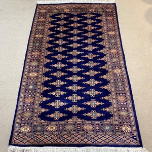 Op de afbeelding: Een blauw tapijt met een traditioneel Perzisch ontwerp. Het tapijt heeft een donkerblauwe achtergrond met een herhalend patroon van gouden, groene en rode bloemmotieven. Het tapijt heeft een franje rand.