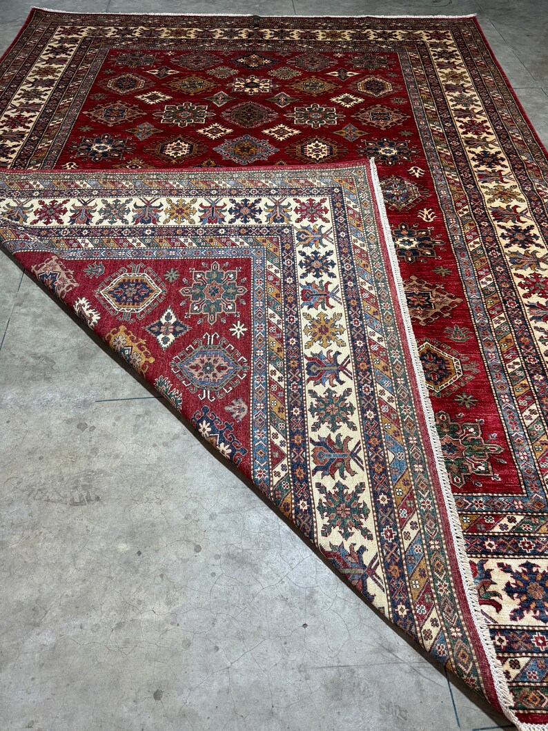 May include: A red, cream, and blue patterned rug with intricate designs. The rug is folded in half, revealing the underside of the rug.