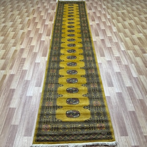 May include: A long, narrow, gold and black patterned rug with a fringed edge. The rug has a traditional design with intricate details.