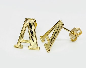 May include: A pair of gold-colored letter "A" stud earrings. The earrings are in a serif font, with a textured surface. The posts and backs are also gold-colored. The earrings are displayed on a white background.