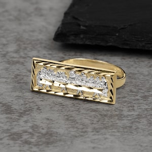 Könnte beinhalten: Ein goldfarbener Ring mit einer rechteckigen Platte, die ein silberfarbenes Relief einer Gruppe von Figuren an einem langen Tisch zeigt. Die rechteckige Platte hat einen texturierten, diamantgeschliffenen Rand und die Ringschiene ist glatt poliert.