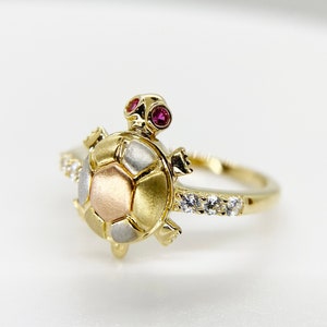 Puede incluir: Un anillo de oro con un diseño de tortuga de tres tonos. El caparazón de la tortuga es de oro, plata y oro rosa. La tortuga tiene dos pequeñas piedras preciosas rojas para los ojos y una banda de pequeñas piedras preciosas transparentes alrededor del anillo.