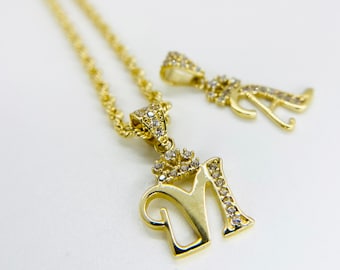 Puede incluir: Collar de cadena de oro con un colgante con la letra 'M' con una corona de pequeñas piedras transparentes. También se incluye un colgante más pequeño con la letra 'A'.