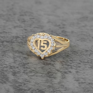 May include: A gold-colored ring featuring a heart-shaped design with the number "15" in the center. The heart is surrounded by a halo of small, clear gemstones. The ring has a split shank design.