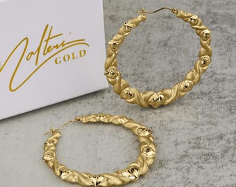 Könnte beinhalten: Ein Paar goldene Creolen mit einem gedrehten Design und diamantförmigen Akzenten. Die Ohrringe liegen auf einer weißen Oberfläche mit einer weißen Box im Hintergrund, auf der "Joltini GOLD" steht.