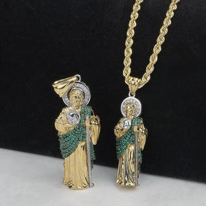 Puede incluir: Dos colgantes dorados que representan una figura religiosa, uno en una cadena de cuerda dorada. Los colgantes presentan un halo, detalles verdes y un báculo. La figura lleva una túnica dorada. El fondo es de color oscuro.