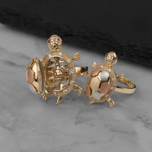 May include: Two gold-toned rings shaped like turtles. One ring features an open shell design revealing a smaller turtle inside, accented with clear stones and red eyes. The second ring has a closed, multi-colored shell and red eyes.