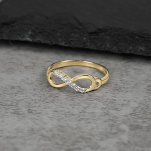 May include: A gold ring featuring an infinity symbol design. One side of the infinity loop is set with a row of small, clear, round-cut stones that sparkle against the polished gold band. The ring has a smooth, continuous metal finish.