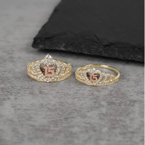 May include: Two gold-toned rings, one larger than the other, each shaped like a crown. The crowns feature a pinkish-gold number "15" at the center, surrounded by clear gemstones. The rings are set against a gray surface.