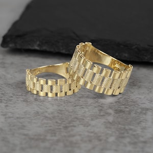 Puede incluir: Dos anillos dorados con un diseño texturizado tipo correa de reloj. Los anillos se muestran sobre una superficie gris con un fondo oscuro y texturizado. Los anillos están hechos de un metal amarillo brillante.