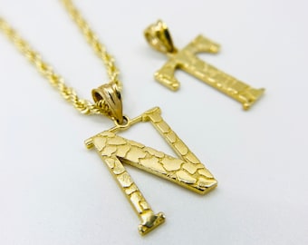 Puede incluir: Collar de cadena de oro con dos colgantes, uno es una letra 'N' y el otro es una cruz. Ambos colgantes tienen una superficie texturizada.