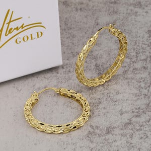 May include: Two gold-coloured hoop earrings with an intricate openwork design. The earrings are displayed on a neutral-toned surface, with a white box partially visible in the background. The box has gold text that reads "Hemi GOLD".