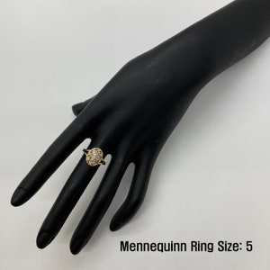 A gold ring with a flower-like cluster of stones has a thin, delicate band and a raised circular setting. The blend of modern and classic styles highlights elegance and sophistication. The size shown is "5" on the mannequin hand.