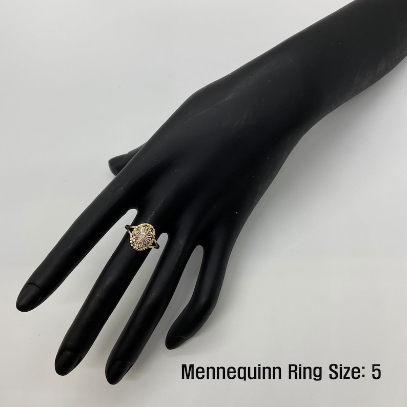 A gold ring with a flower-like cluster of stones has a thin, delicate band and a raised circular setting. The blend of modern and classic styles highlights elegance and sophistication. The size shown is "5" on the mannequin hand.
