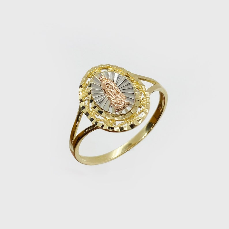A delicate gold ring features an oval-shaped design. The center showcases an image of the Virgin Mary in rose gold, surrounded by a sunburst pattern in yellow gold. The ring's band is thin and slender, crafted in white gold.