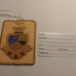 May include: A tan luggage tag with a gold embroidered crest featuring a crown, a blue and gold shield, and the letters "H.O.P.B."  The tag has a white label with the words "Name", "Address", "City", "State", "Zip Code", and "Telephone".