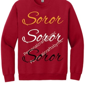 Peut inclure: Un sweat-shirt rouge avec le mot "Soror" en trois couleurs : or, blanc et noir. Le texte "PersonalcraftsbyMC" est imprimé sous le texte blanc et noir.
